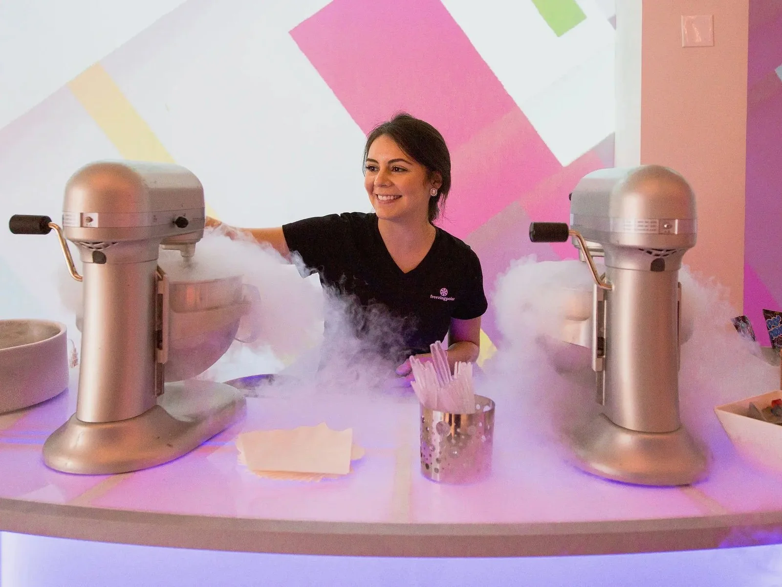 Smiling server between two fogging mixers at a purple-lit Glow Bar for liquid nitrogen ice cream catering Houston
