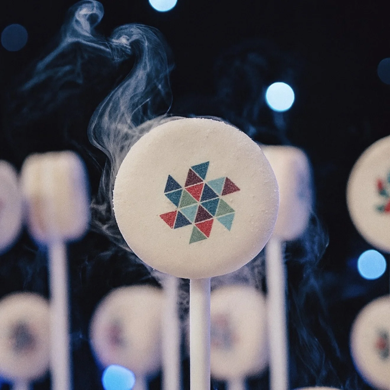 Close-up of a smoking NitroPop Macaron printed with a colorful geometric client logo, with more logo macarons and blue lights behind it at a corporate event